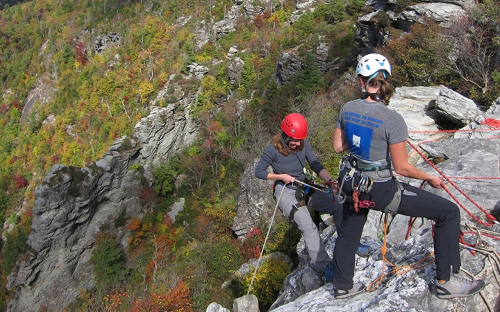 Two people wearing safety gear are secured by ropes near the edge of a cliff. One person appears to be an instructor, giving direction to the other person. Behind them is a rocky landscape covered in greenery. 